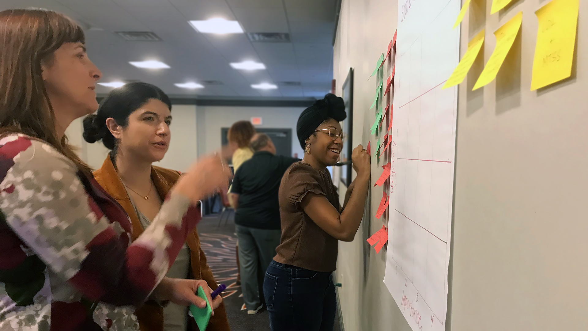 Photo of two adult women engaged in discussion pointing at a wall with sticky notes and chart paper. A woman in the background is smiling and writing on a sticky note on the wall.